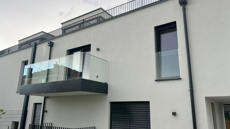 Moderne Gebäudefassade mit einem eleganten Glasbalkon und einer Terrasse im Erdgeschoss.