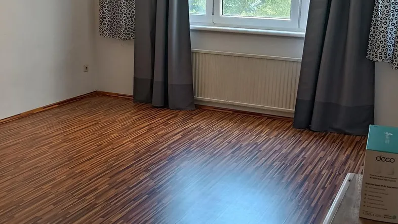 Heller Raum mit Holzstreifenboden, großen Fenstern und Blick ins Grüne, ausgestattet mit Vorhängen.