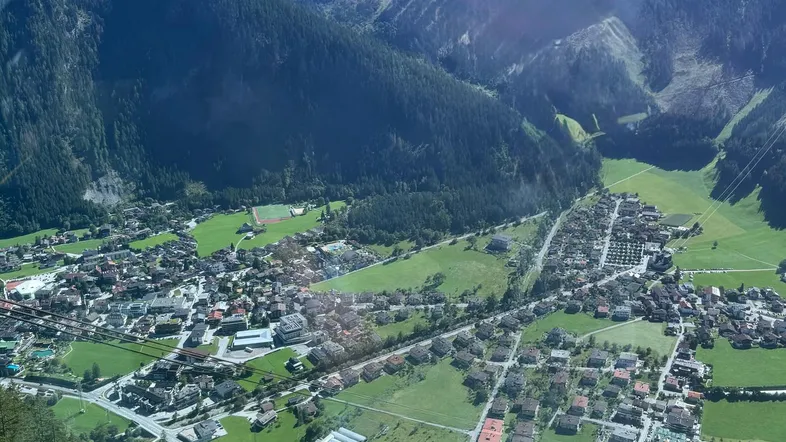 Luftaufnahme eines malerischen Tals mit einem Dorf, umgeben von majestätischen Bergen und dichten Wäldern unter blauem Himmel.