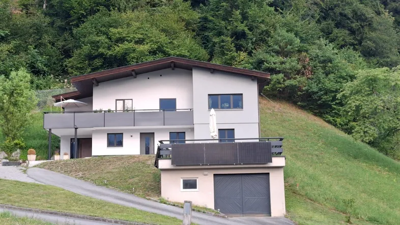 Modernes Haus am Hang mit Balkonen und integrierter Garage, umgeben von grünen Bäumen.