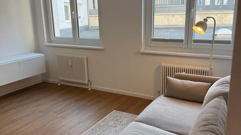 Helles Wohnzimmer mit modernem Sofa, Holzboden und großen Fenstern, die viel Tageslicht hereinlassen.