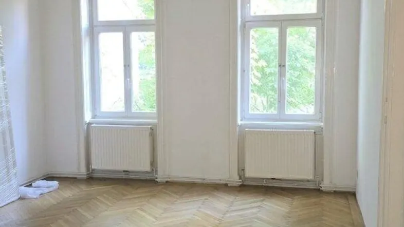 Helles Zimmer mit Fischgrätparkett und zwei großen Fenstern, das viel Tageslicht hereinlässt.