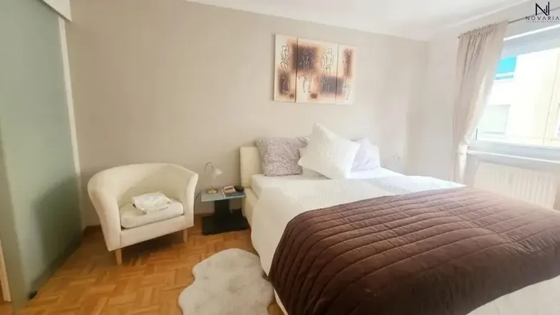 Helles Schlafzimmer mit Parkettboden, beigem Sessel und einem großen Bett.
