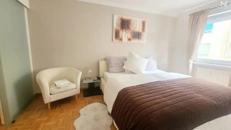 Helles Schlafzimmer mit Parkettboden, Doppelbett, beigen Wänden und einem Fenster mit Vorhängen.