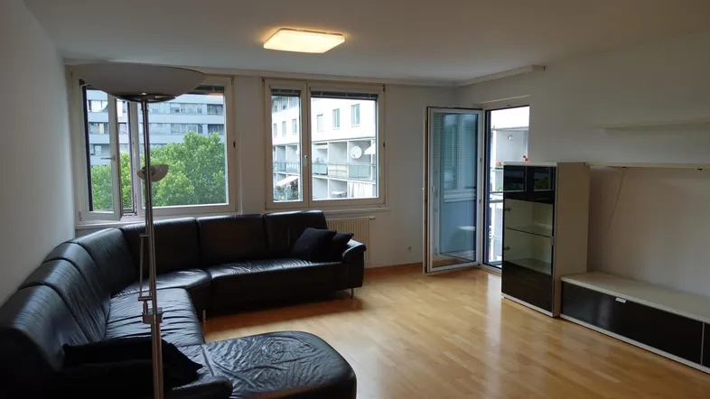 Helles Wohnzimmer mit Parkettboden, schwarzem Ledersofa und Zugang zum Balkon.