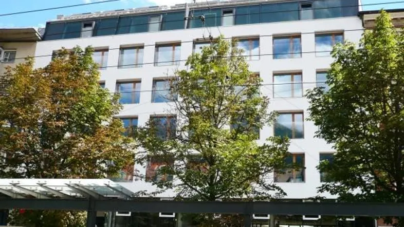 Modernes Gebäude mit vielen Fenstern und Bäumen davor in städtischer Umgebung.