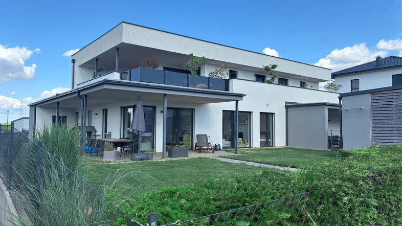 Modernes Gebäude mit großer Terrasse, Garten und Obergeschoss-Balkon unter blauem Himmel.