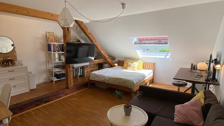 Helles Dachgeschosszimmer mit Holzbalken, Schlafbereich, Wohnbereich und Arbeitsplatz unter einem Dachfenster.