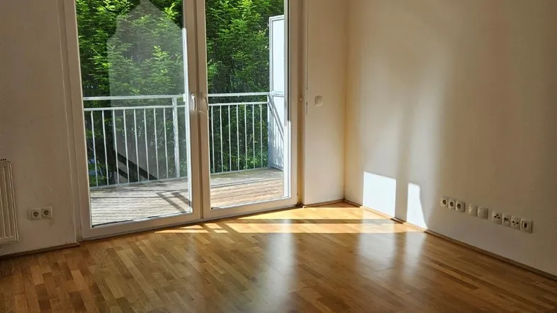 Helles Zimmer mit Holzboden und Zugang zu einem Balkon mit Blick ins Grüne.