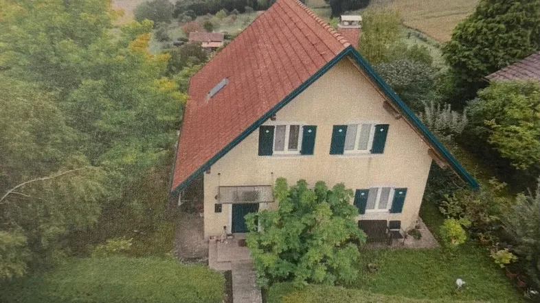 Haus mit rotem Ziegeldach und grünen Fensterläden, umgeben von üppiger Vegetation.