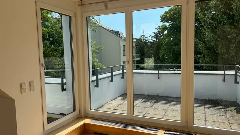 Helles Zimmer mit großen Fenstern und Zugang zu einer sonnigen Terrasse mit Blick ins Grüne.