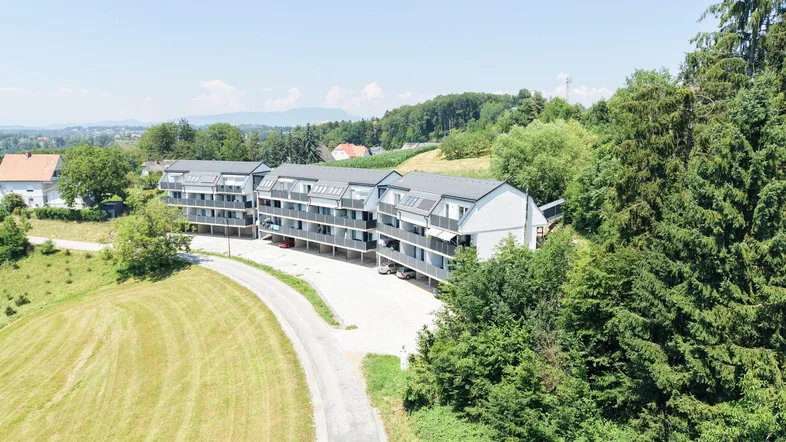 Moderne Wohnanlage mit mehreren Gebäuden, umgeben von grünen Hügeln und Bäumen unter blauem Himmel.