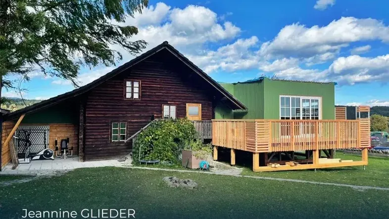 Blockhaus mit grünem Anbau und Holzterrasse unter blauem Himmel, umgeben von Natur.
