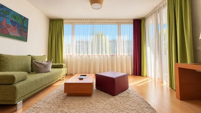 Helles Wohnzimmer mit beigem Teppich, grünem Sofa und großen Fenstern mit Vorhängen.