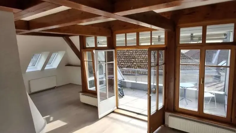 Helles Wohnzimmer mit sichtbaren Holzbalken, Kamin und Zugang zu einer sonnigen Terrasse.