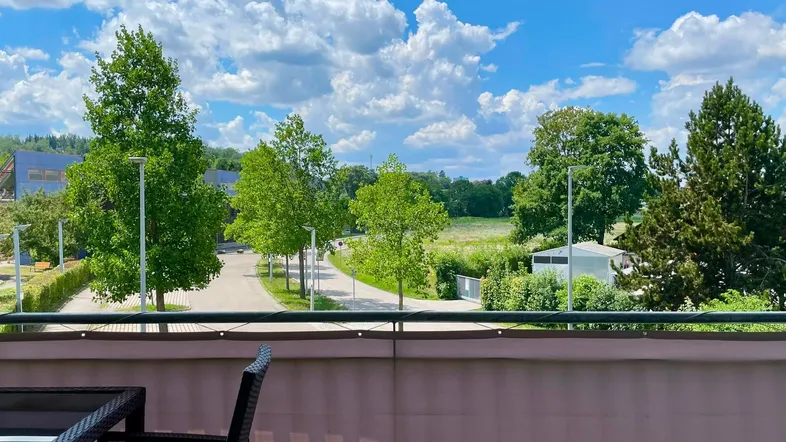 Blick vom Balkon auf eine grüne Umgebung mit Bäumen und einer ruhigen Straße unter blauem Himmel.