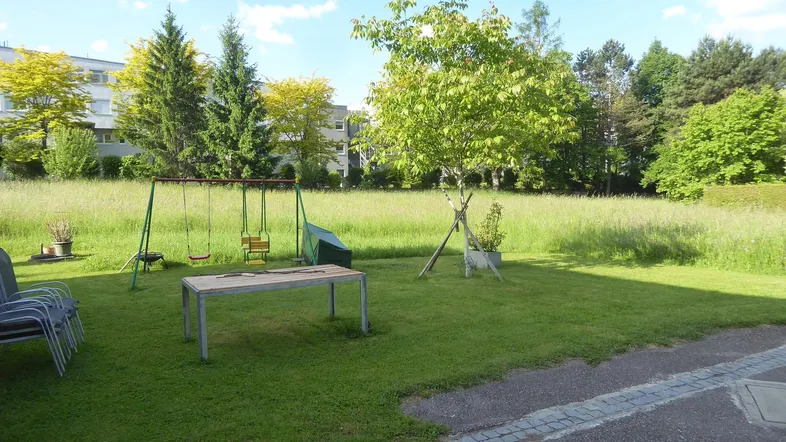 Grüne Außenanlage mit Spielplatz und Rasenfläche, umgeben von Bäumen und Wohngebäuden.