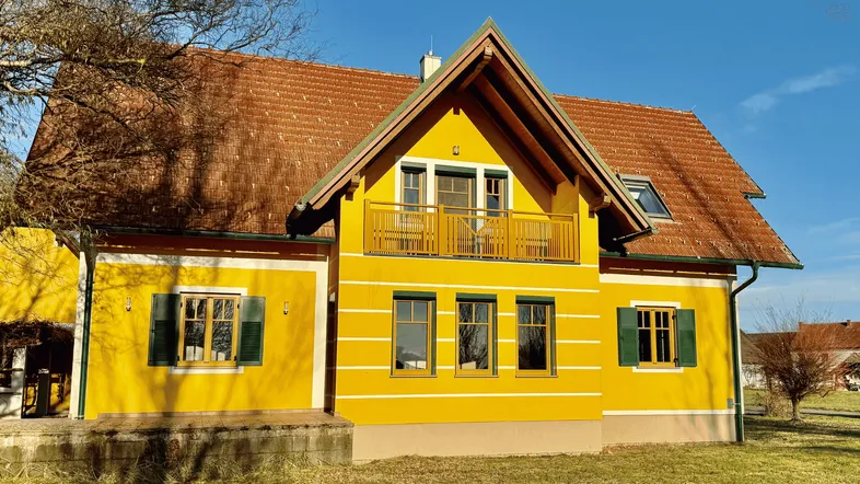 Gelbes Haus mit rotem Ziegeldach und einem Balkon im Obergeschoss. Grüne Fensterläden ergänzen die Fassade.