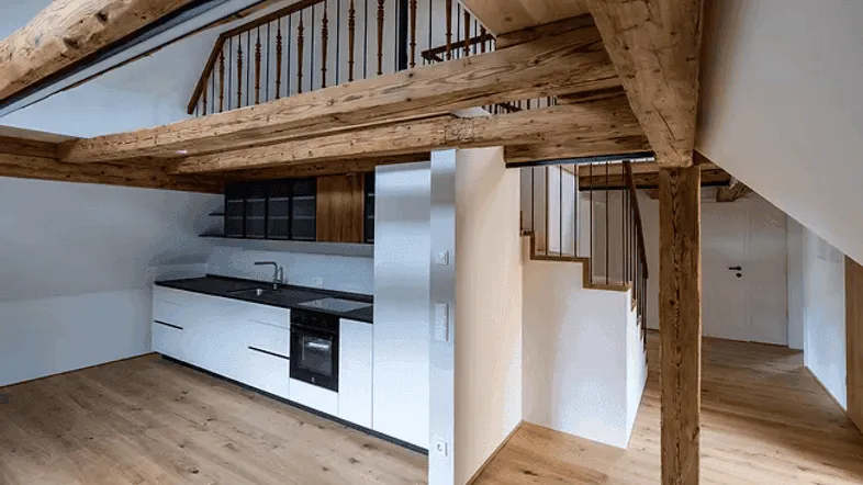 Moderne Küche mit weißen Fronten, dunkler Arbeitsplatte und sichtbaren Holzbalken in einem offenen Raum.