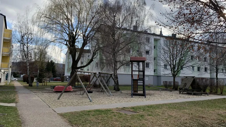 Spielplatz mit Schaukeln und Klettergerüst vor einem Mehrfamilienhaus mit kahlen Bäumen.