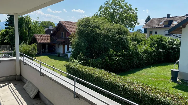 Geräumiger Balkon mit Fliesenboden und Blick ins Grüne, umgeben von Hecken und Bäumen.