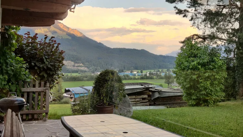 Gemütliche Terrasse mit Esstisch und Grill, umgeben von grüner Landschaft und Bergblick bei Sonnenuntergang.