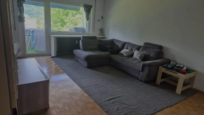 Gemütliches Wohnzimmer mit großem Ecksofa, Parkettboden und Zugang zum Balkon mit grüner Aussicht.