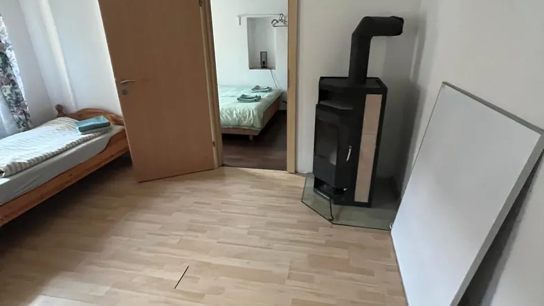 Helles Zimmer mit Holzboden, einem Einzelbett und einem gemütlichen Holzofen, der zu einem weiteren Schlafzimmer führt.