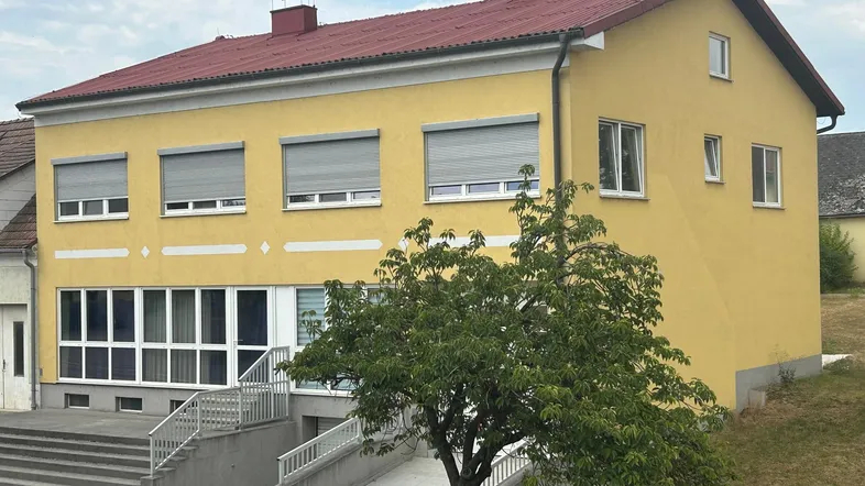 Gelbes Gebäude mit rotem Satteldach, vielen Fenstern und Rollläden, umgeben von Grünflächen.