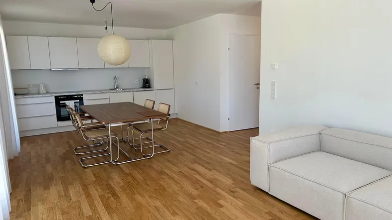 Heller offener Wohn-Essbereich mit moderner Einbauküche, Holzboden und beiger Couch.