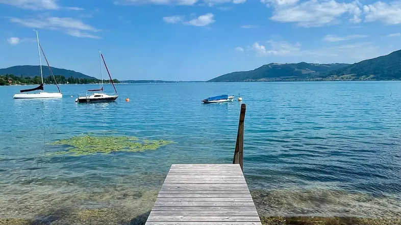Langer Holzsteg führt ins klare Seewasser mit Segelbooten und Bergpanorama unter blauem Himmel.