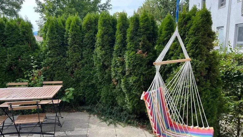 Sonniger Terrassenbereich mit Esstisch und einem bunten Hängesessel, umgeben von hohen Hecken.