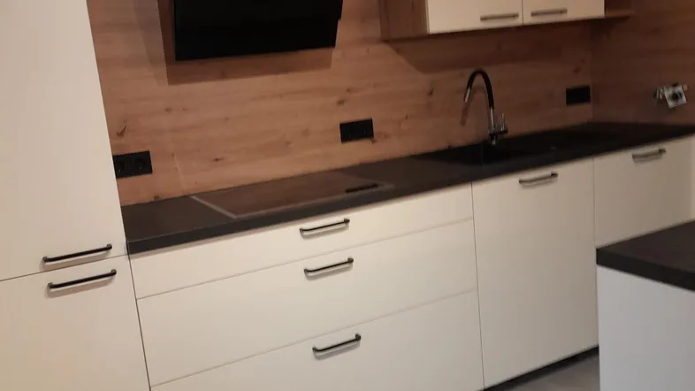 Moderne Küche mit weißen Schränken, Holzrückwand, schwarzer Arbeitsplatte und Induktionskochfeld.