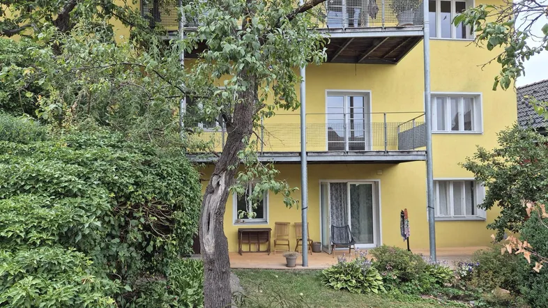 Gelbe Fassade eines Mehrfamilienhauses mit Balkonen und einer möblierten Terrasse im Erdgeschoss.