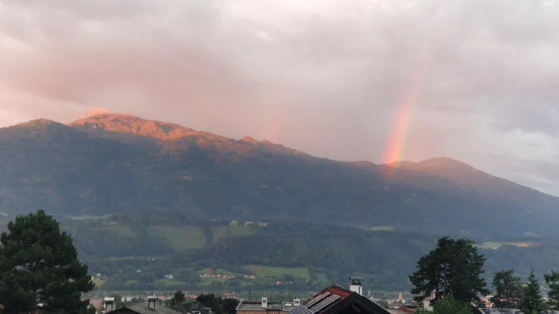 Malerischer Bergblick mit einem Regenbogen über den Gipfeln und Dächern im Vordergrund.