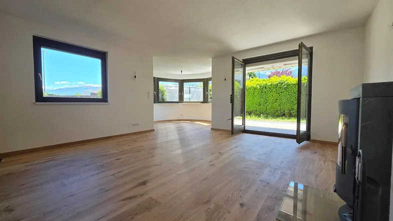 Helles Wohnzimmer mit Holzboden, Kaminofen und Zugang zur Terrasse mit Hecke.