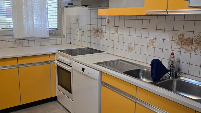 Helle Küche mit gelben Schränken, Fliesenboden, Fenster und integrierten Geräten.