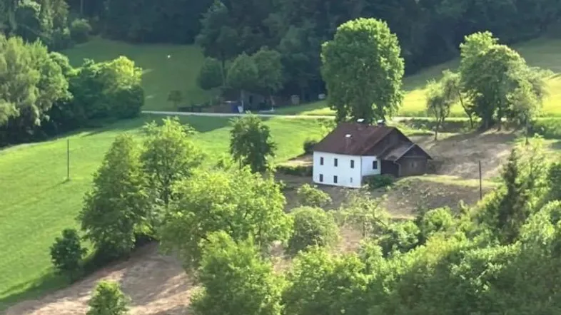 Ländliches Haus eingebettet in eine grüne Hügellandschaft mit Wald im Hintergrund unter blauem Himmel.