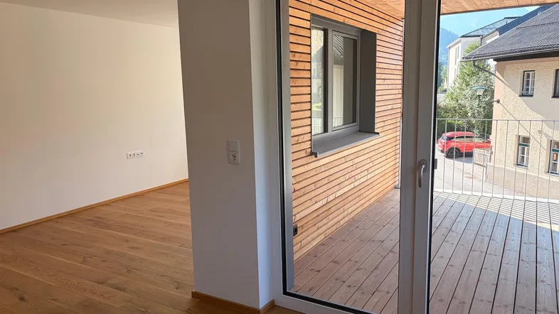 Helles Zimmer mit Holzboden und Zugang zu einem Balkon mit Holzverkleidung.