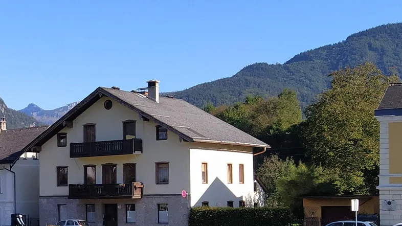 Traditionelles Haus mit Satteldach und Balkonen, umgeben von Bergen und grüner Landschaft.