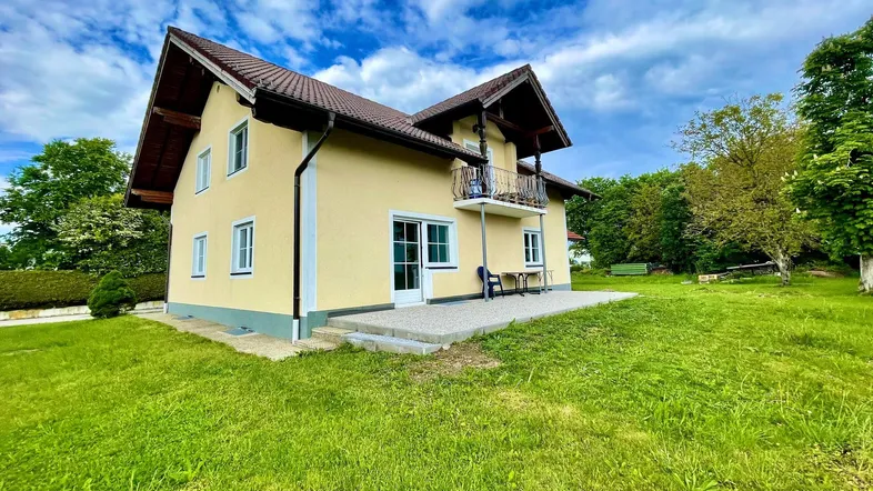 Gelbes Einfamilienhaus mit braunem Dach, umgeben von grünem Rasen und Bäumen unter blauem Himmel.