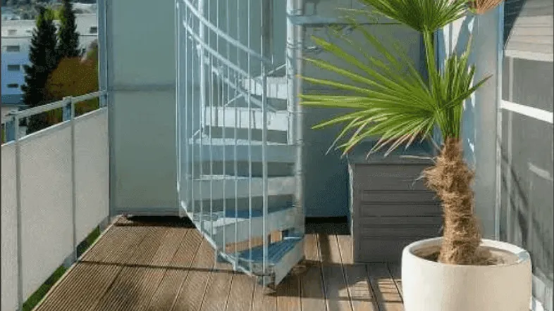 Geräumiger Balkon mit Holzfußboden, Wendeltreppe und Topfpflanzen unter blauem Himmel.