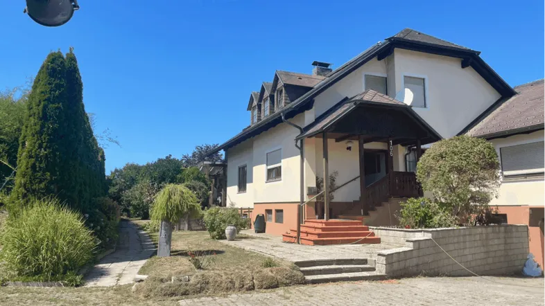 Ansicht des Hauses mit gepflastertem Vorplatz, Treppe zum Eingang und umliegender Vegetation.