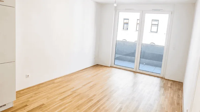 Helles, leeres Zimmer mit Parkettboden und Zugang zu einem Balkon, ideal als Wohnbereich.