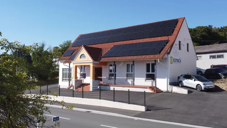 Modernes Gebäude mit Photovoltaikanlage auf dem Dach und Parkplätzen davor, umgeben von Grün.