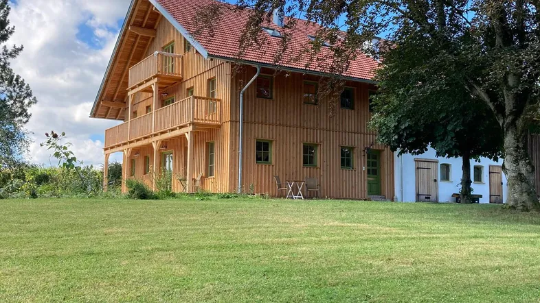 Holzhaus mit rotem Ziegeldach und Balkonen, umgeben von einem grünen Rasen unter blauem Himmel.