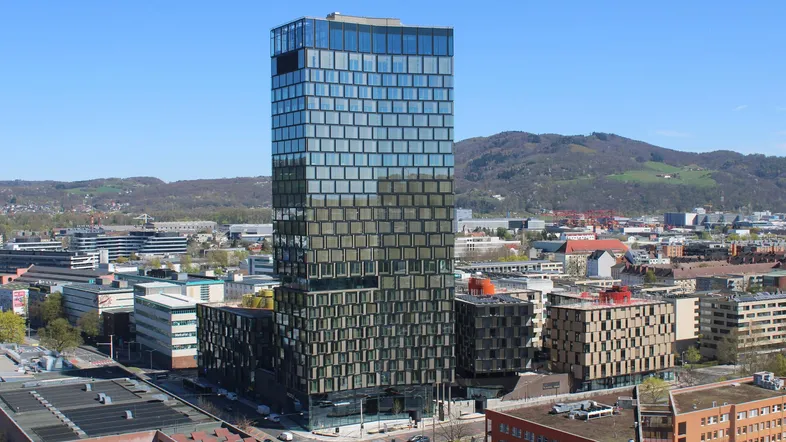 Hohes modernes Gebäude mit Glasfassade, umgeben von städtischer Bebauung und grünen Hügeln im Hintergrund.