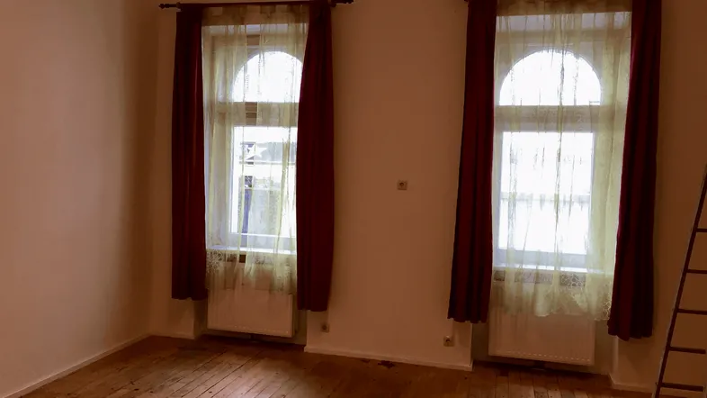 Heller Raum mit zwei hohen Rundbogenfenstern, Holzfußboden und roten Vorhängen.