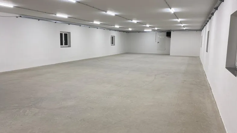 Großer, leerer Raum mit weißen Wänden, Betonboden und mehreren kleinen Fenstern, beleuchtet von Deckenleuchten.