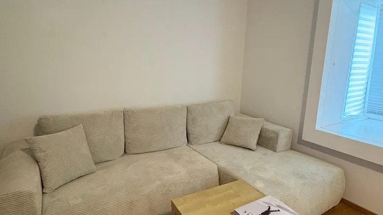 Gemütliches Wohnzimmer mit beigem Ecksofa, Holztisch und hellem Fensterbereich.